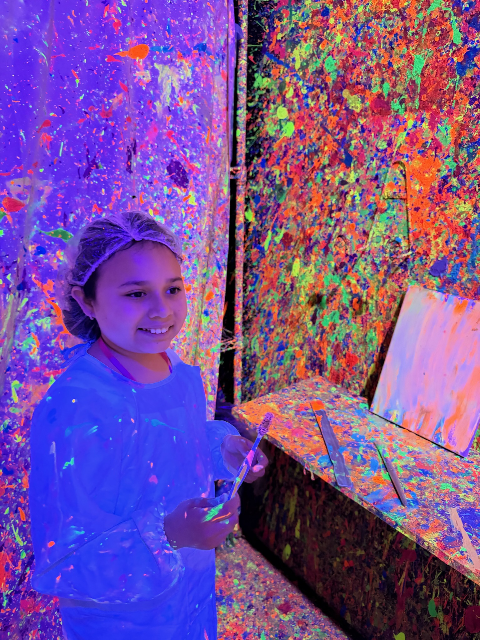 Young girl smiling in white smock holding a paintbrush in the Paint Me One blacklight room
