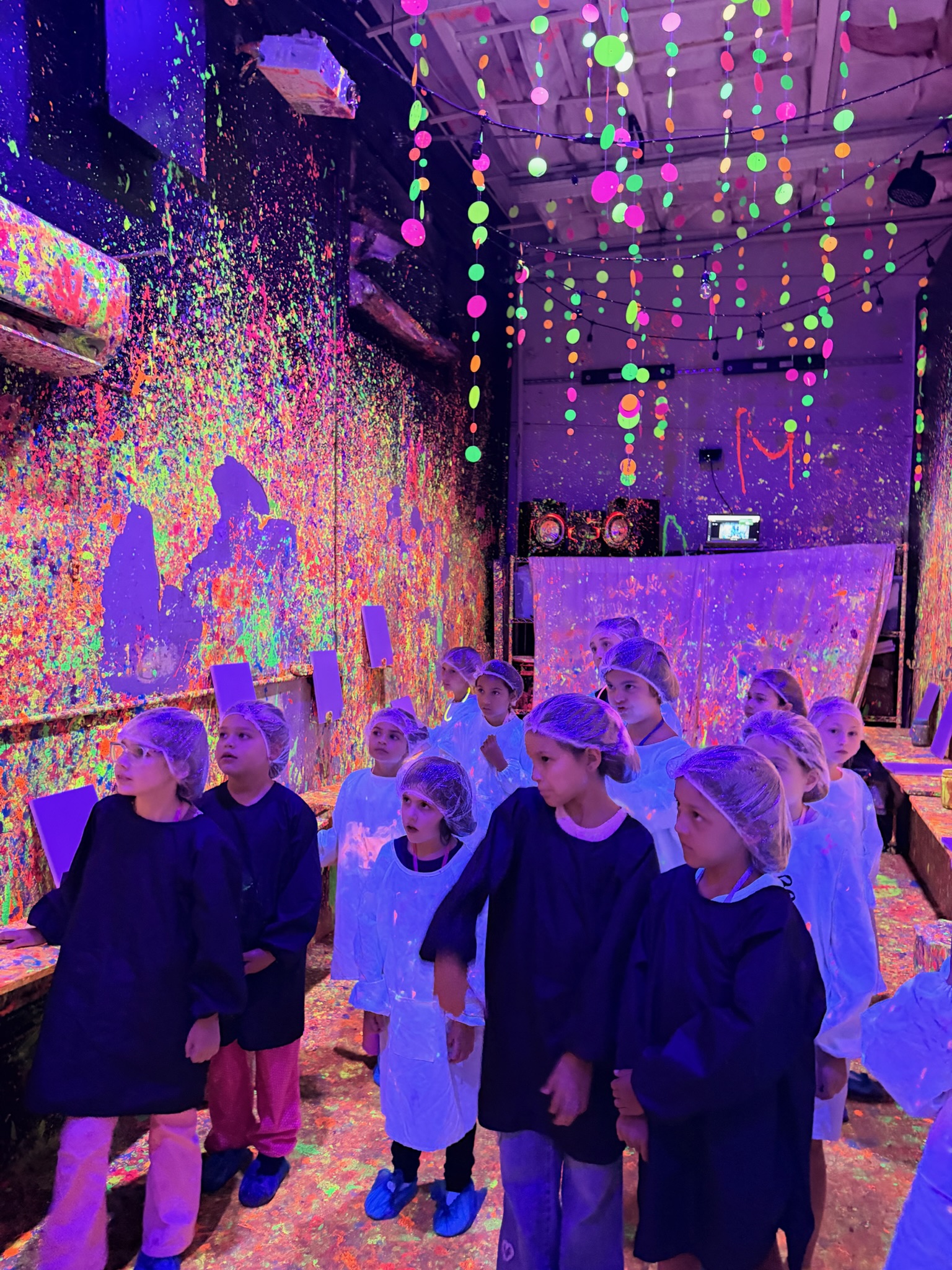 Large group of kids in black smocks and hair nets standing in the neon blacklight Paint Me One room