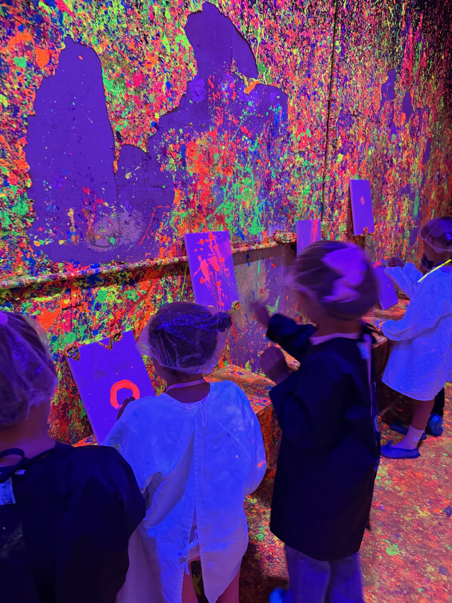 Kids in white lab coats painting glowing canvases against the neon splatter wall