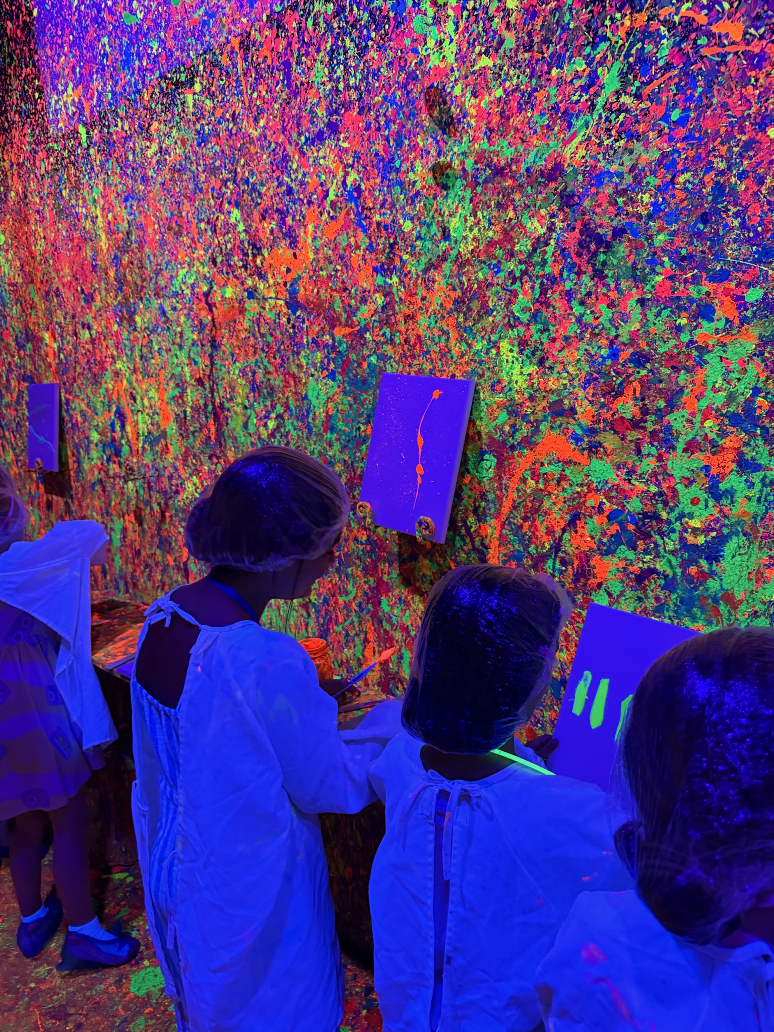 Kids in white lab coats painting canvases surrounded by glowing neon walls